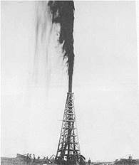 Black-and-white photograph of an oil derrick with a gusher of oil shooting from the top